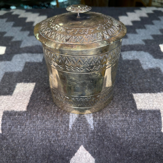 A silver toilet paper holder with intricate hand-tooled designs on the lid and sides, placed on a textured grey background.